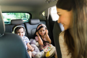 Happy little boy and girl with headphones watching a movie on a tablet while sitting in the car. Fun young woman driving with her cute kids